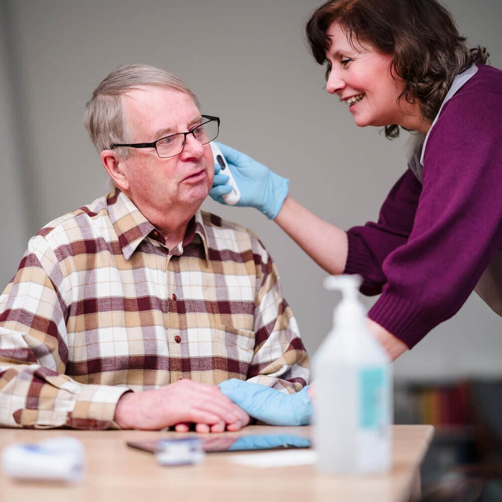 A caregiver wearing gloves checks the ear of an elderly man seated at a table, both smiling. - Home Instead