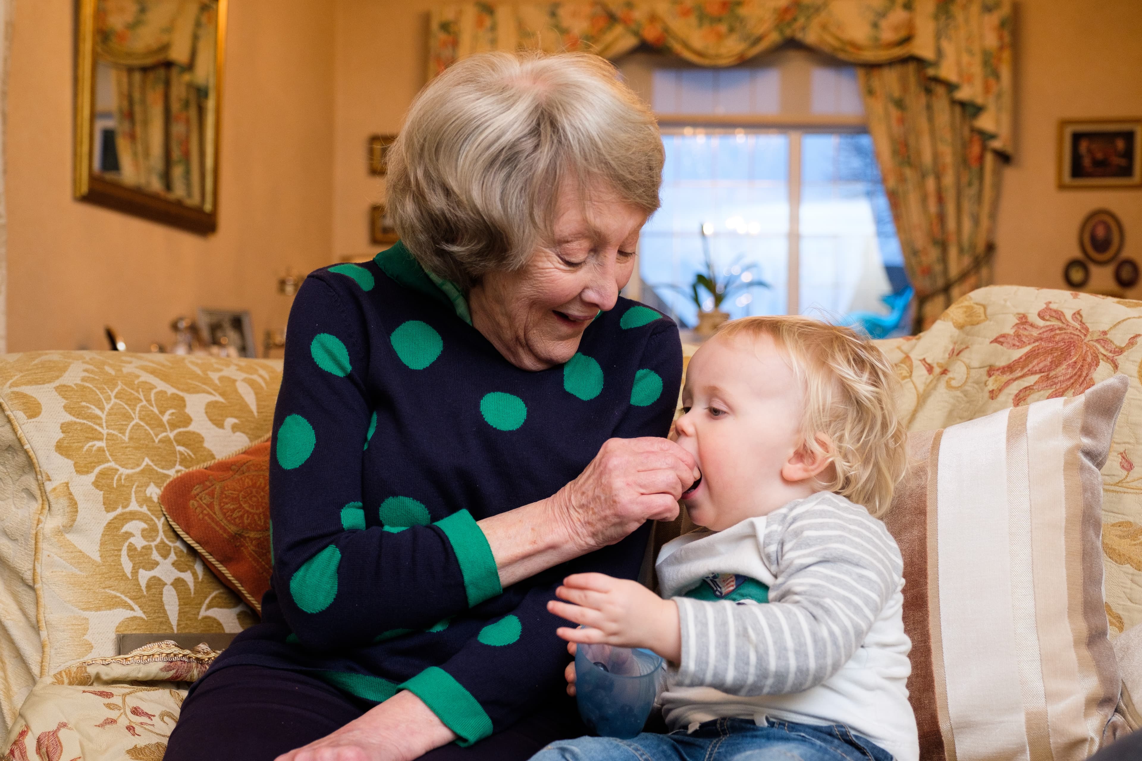 An elderly woman and a toddler share a loving moment on a couch, with the woman feeding the child a snack. - Home Instead