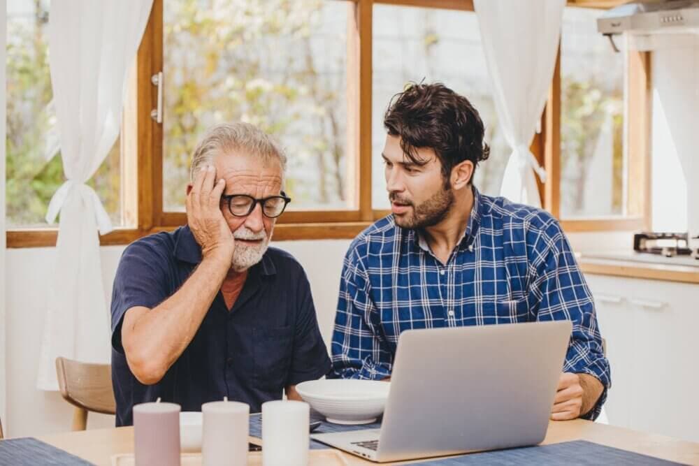 Two men sitting at a table with a laptop, one looking stressed while the other offers assistance. - Home Instead