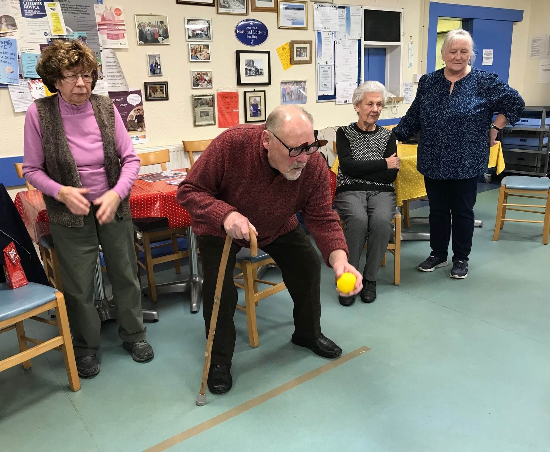 A group of elderly people enjoy an indoor game; a man with a cane is about to throw a yellow ball. - Home Instead