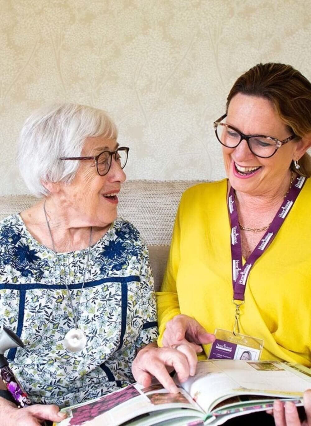 An elderly woman and a younger woman with a nametag are smiling and looking at a book together on a couch. - Home Instead