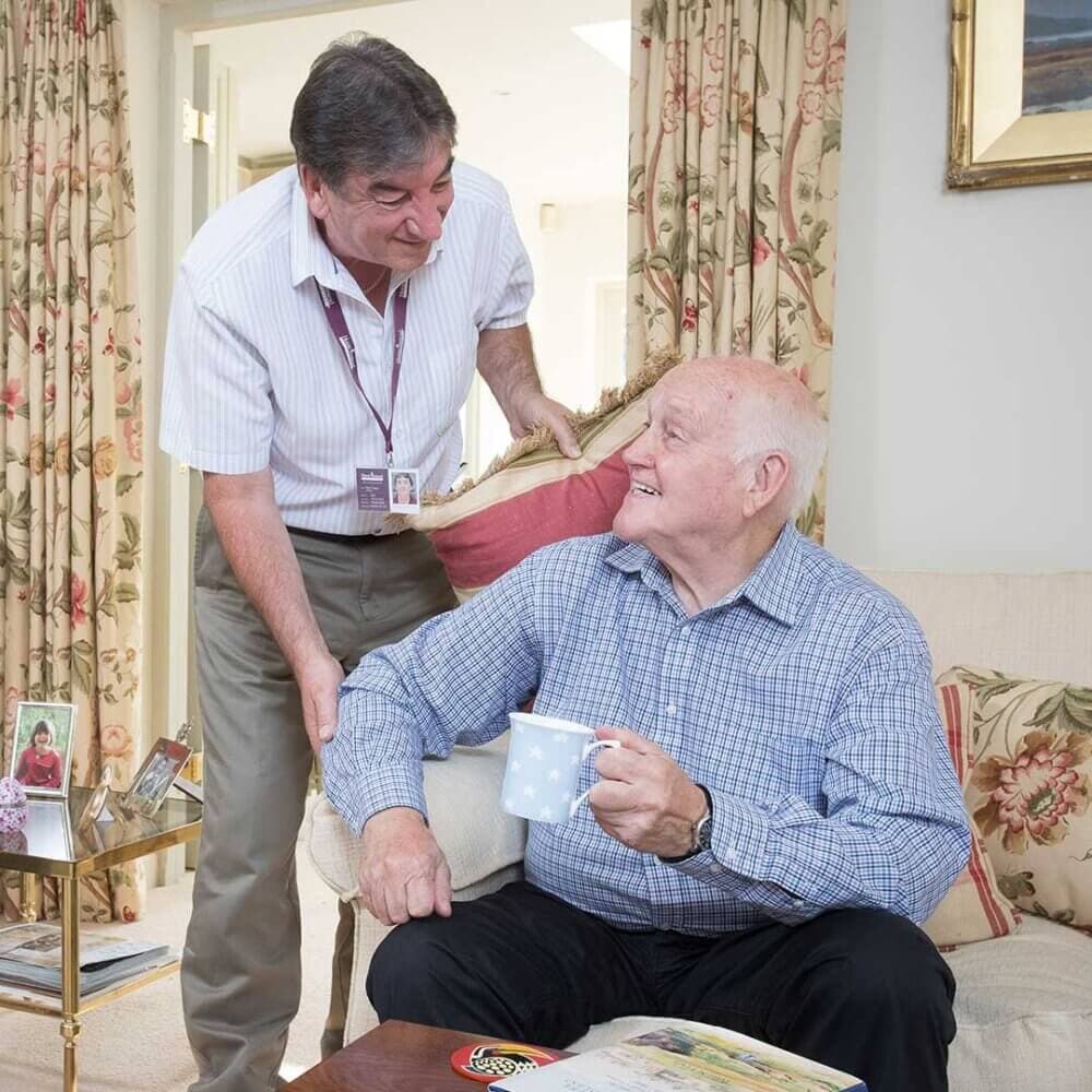 A man stands beside an older adult seated in a living room, both smiling; the older adult holds a mug. - Home Instead