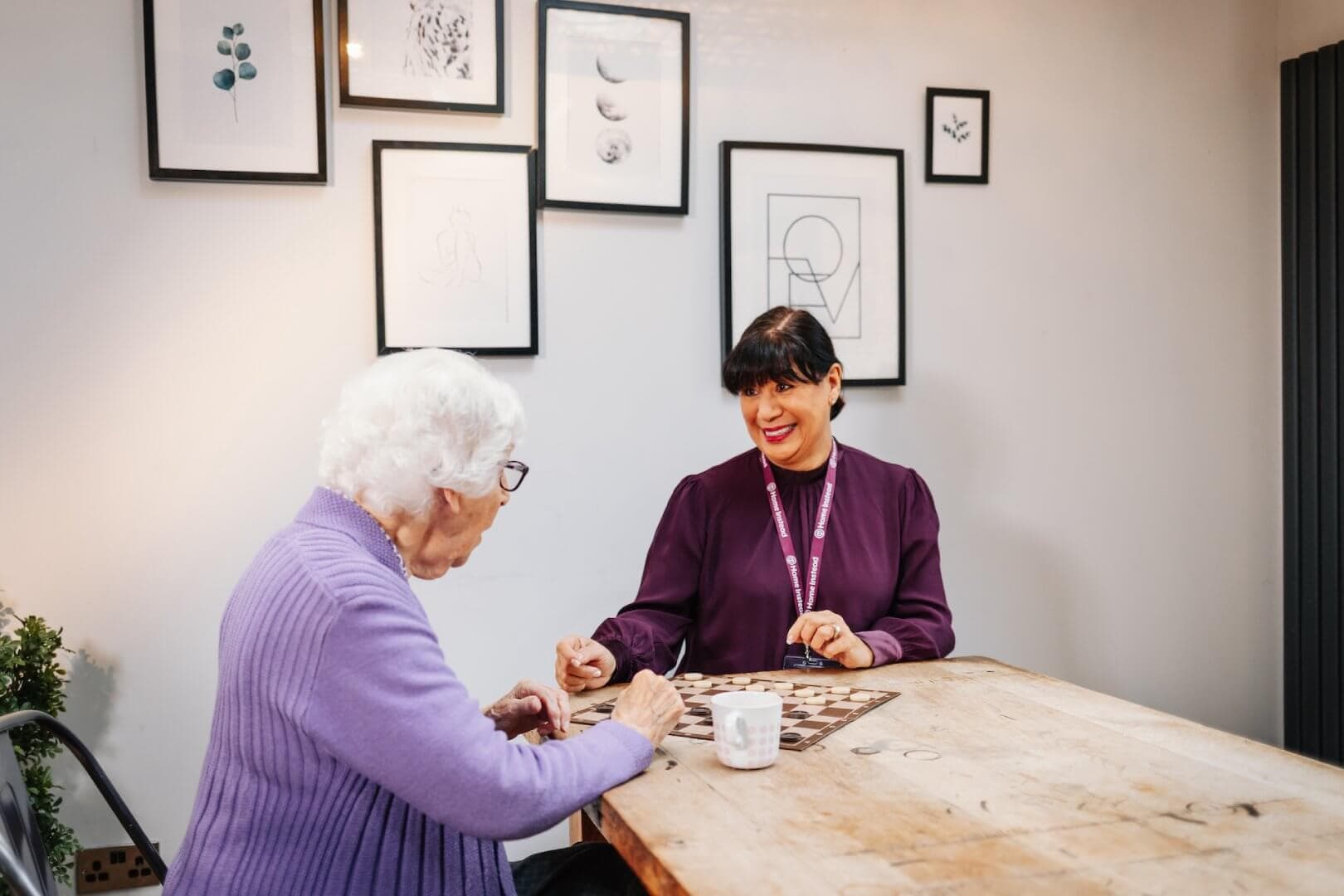 An elderly woman and a caregiver smiling, sitting at a table playing a game in a cozy room with framed art on the walls. - Home Instead