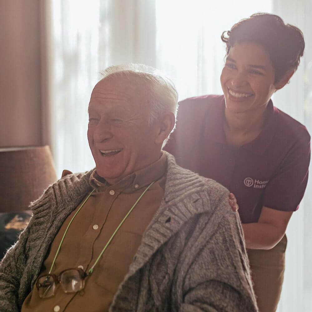 A caregiver smiles with an elderly man in a cozy home setting, both appear happy and in good spirits. - Home Instead