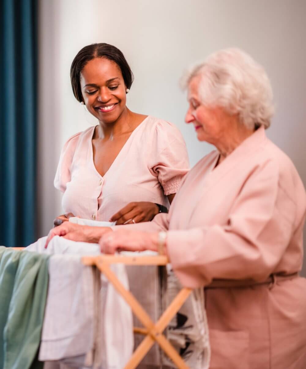 Two women smiling while folding laundry together, one older, one younger, both wearing light pink clothes. - Home Instead