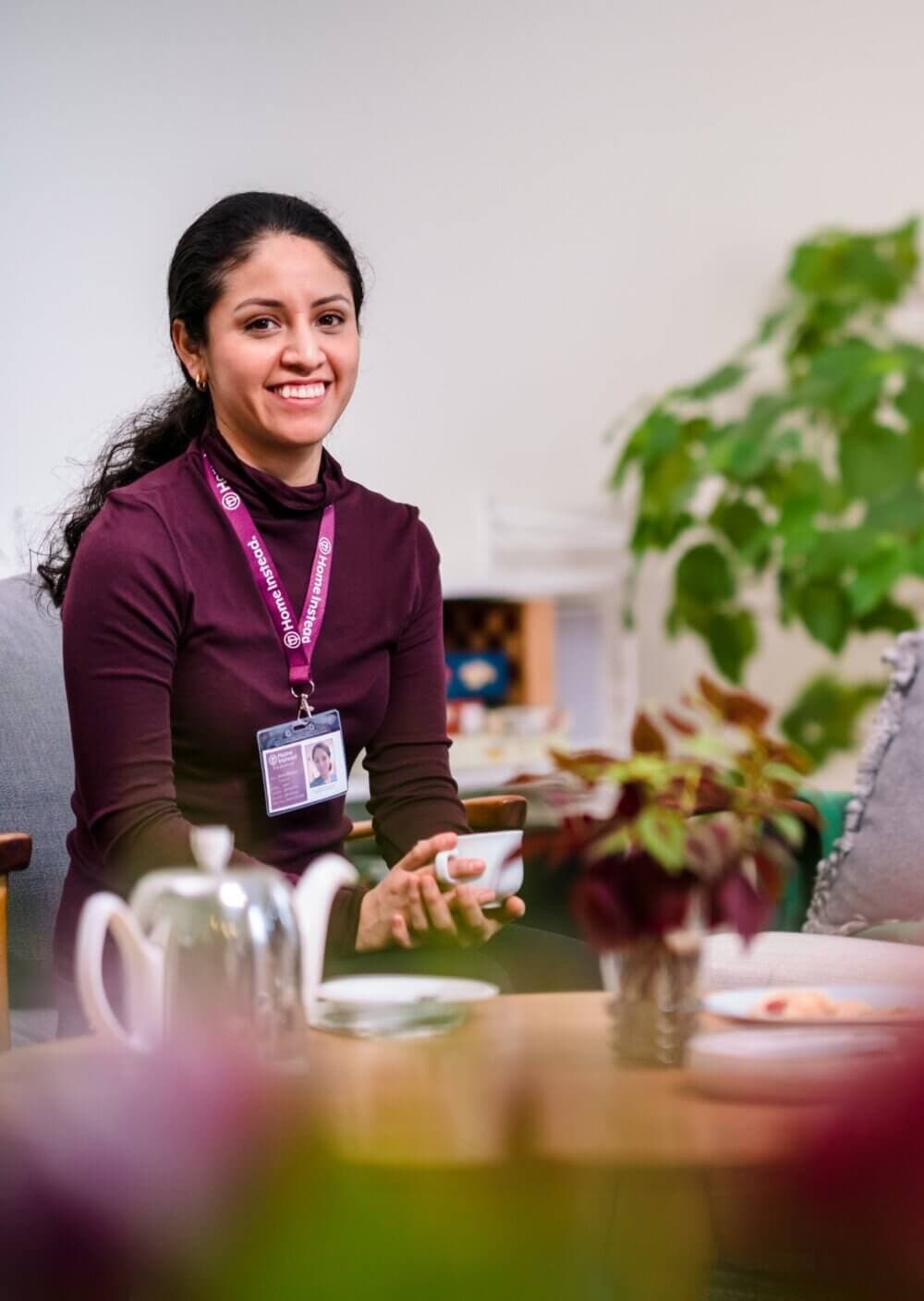 Smiling person with dark hair in a maroon top and ID badge, holding a cup, sitting indoors near a table with a teapot. - Home Instead