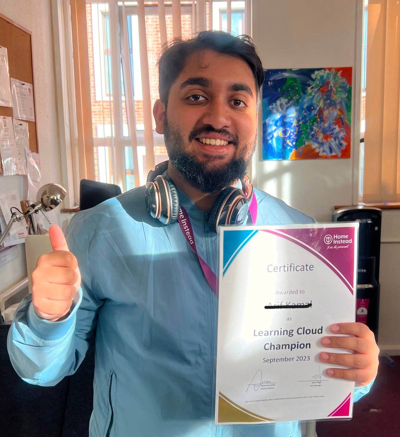 A man with headphones gives a thumbs up and holds a "Learning Cloud Champion" certificate. - Home Instead