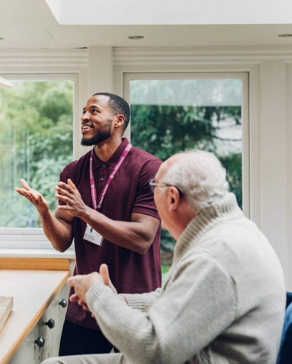 A man in a maroon shirt engages in conversation with an elderly man in a gray sweater in a bright room with windows. - Home Instead