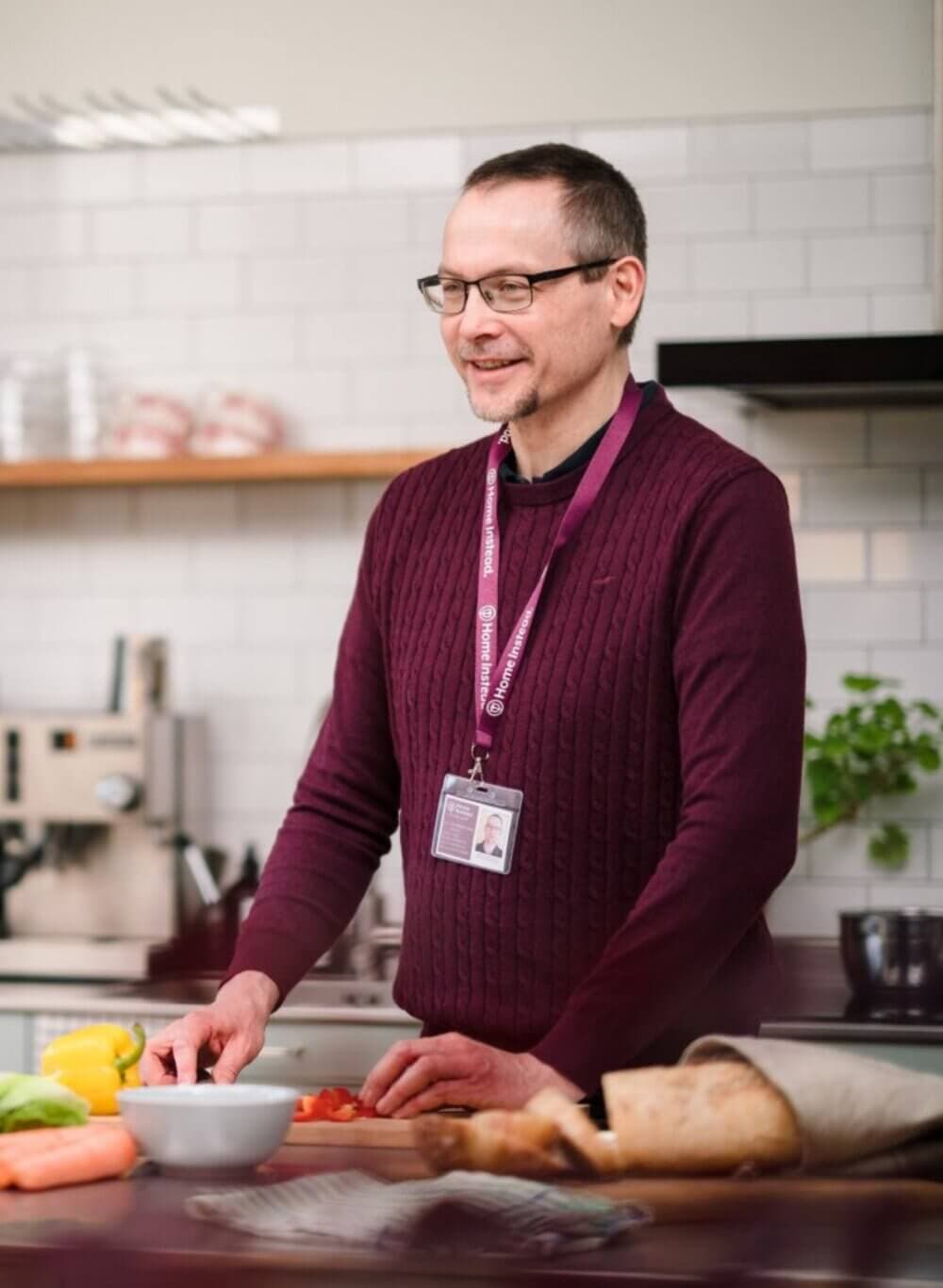 A person wearing glasses and a maroon sweater stands in a kitchen, chopping vegetables on a wooden counter. - Home Instead