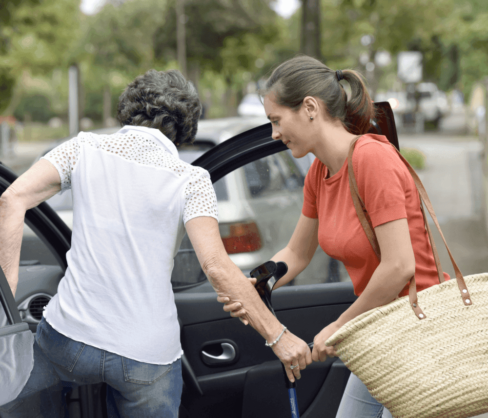 A young woman helps an elderly woman with a cane get into a car on a sunny day. - Home Instead