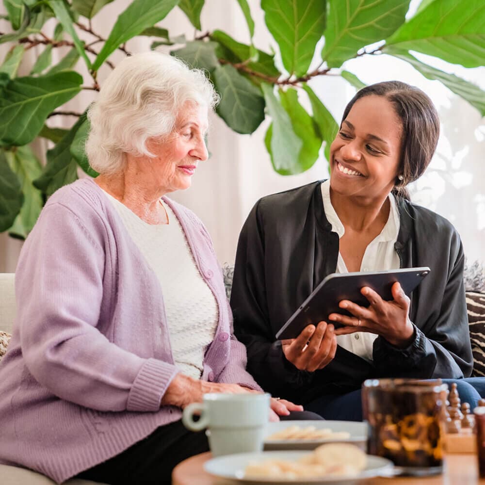 Elderly woman and a young woman smiling while looking at a tablet together, with a potted plant in the background. - Home Instead