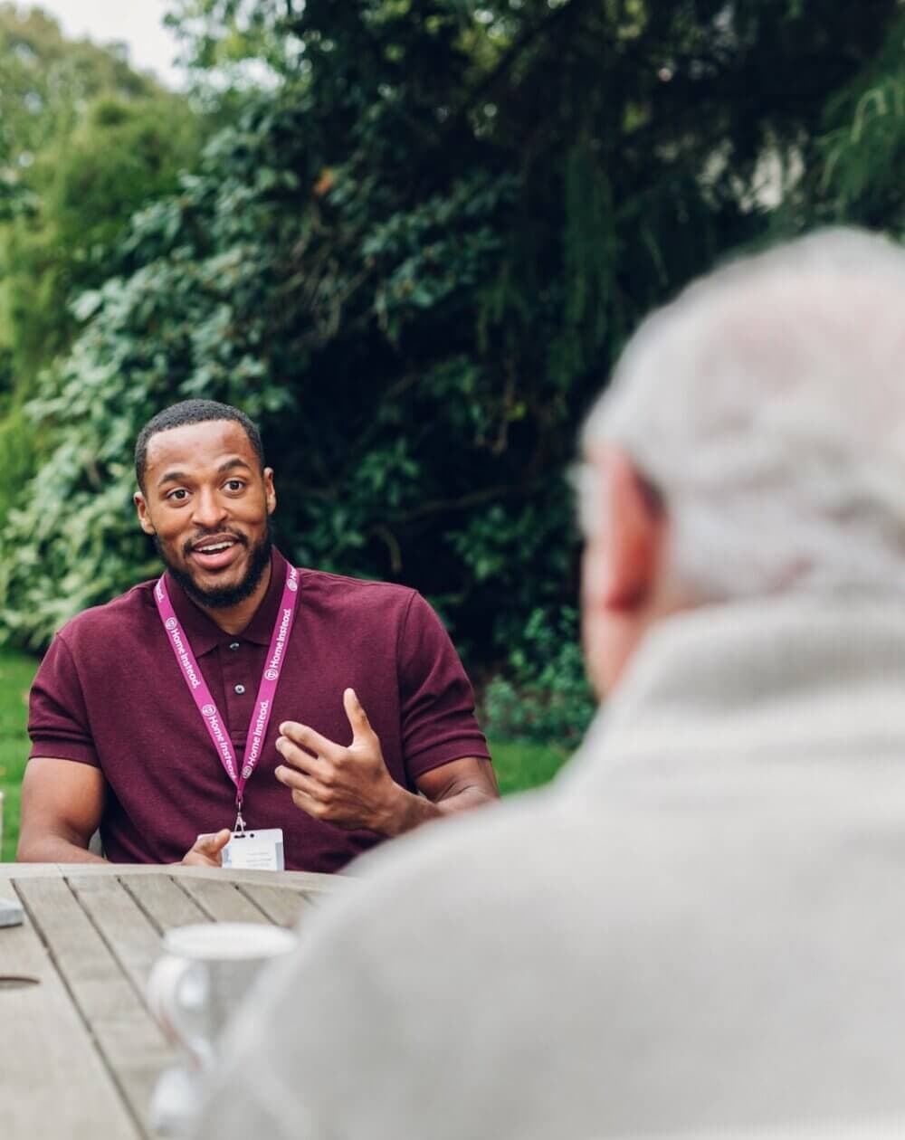An elderly couple sits laughing on a bench around a tree in a green park. - Home Instead