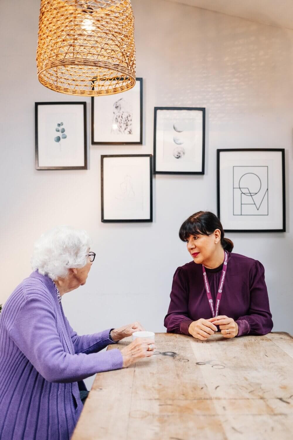 An elderly woman and a younger woman sit at a wooden table, talking. Art frames hang on the wall behind them. - Home Instead