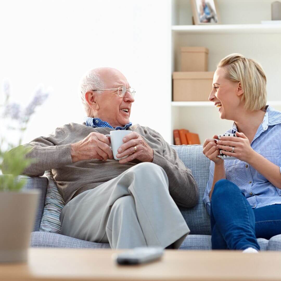 An elderly man and a young woman sit on a couch, smiling and holding mugs, enjoying a conversation. - Home Instead