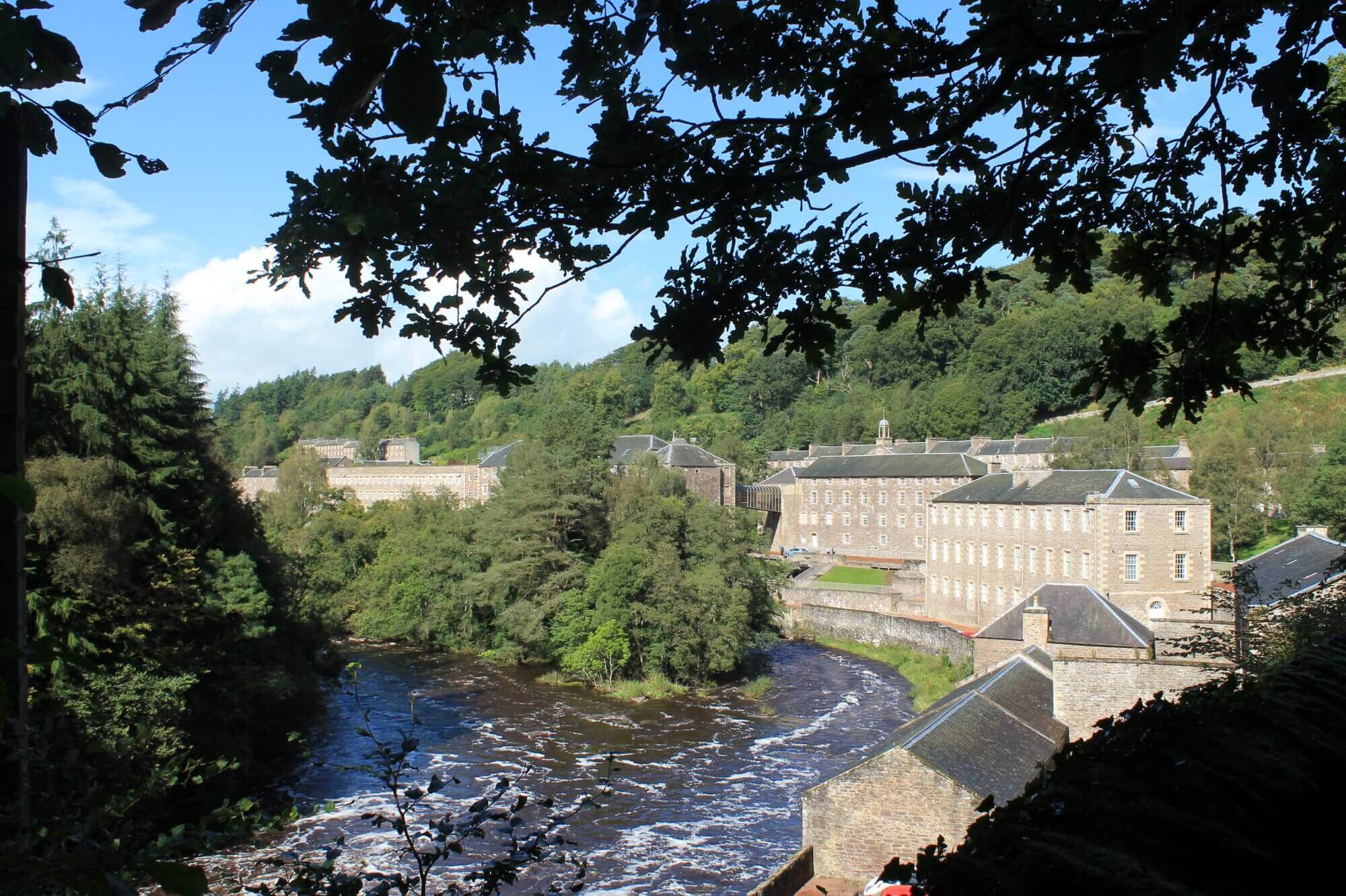 Scenic view of a historic mill complex by a river, surrounded by trees and hills under a partly cloudy sky. - Home Instead