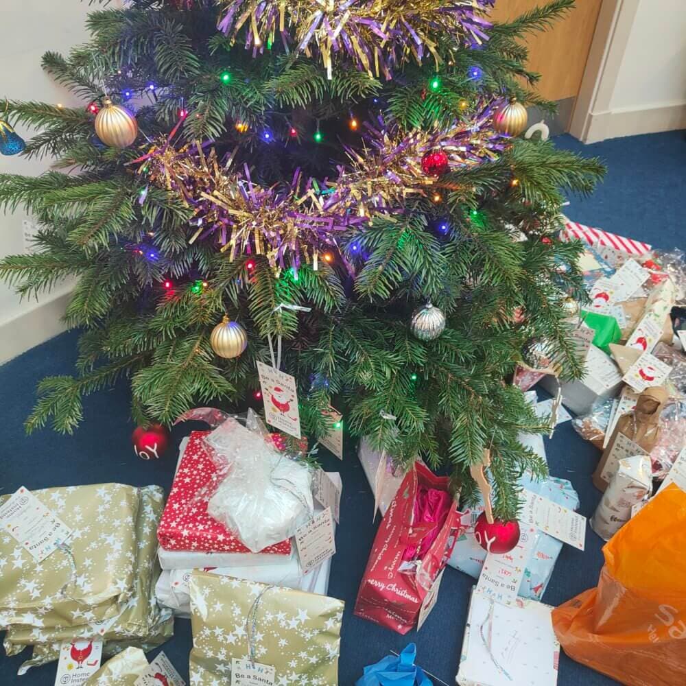 A decorated Christmas tree with colorful lights and tinsel, surrounded by numerous wrapped presents underneath. - Home Instead