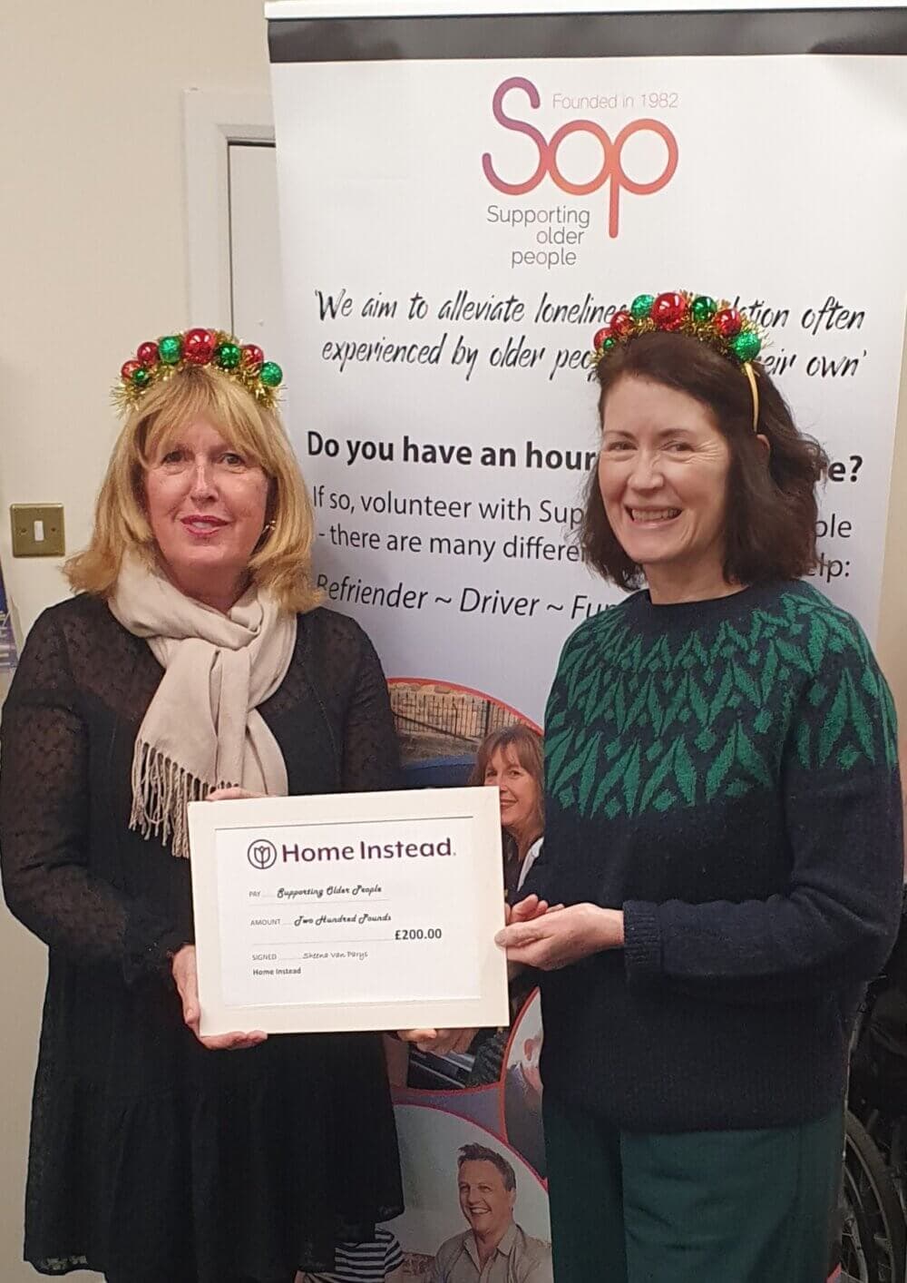 Two women wearing festive headbands hold a framed donation certificate in front of a "Supporting Older People" banner. - Home Instead