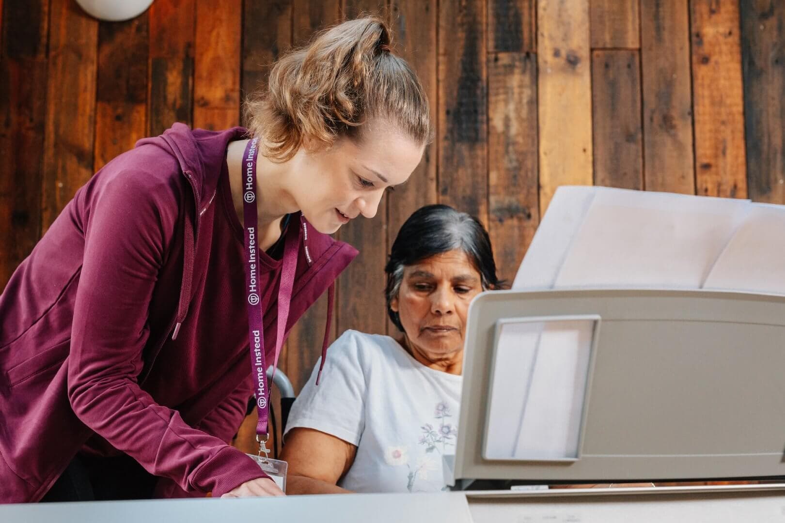 A young woman helps an older woman read or use a device indoors. They appear focused and engaged in the task. - Home Instead