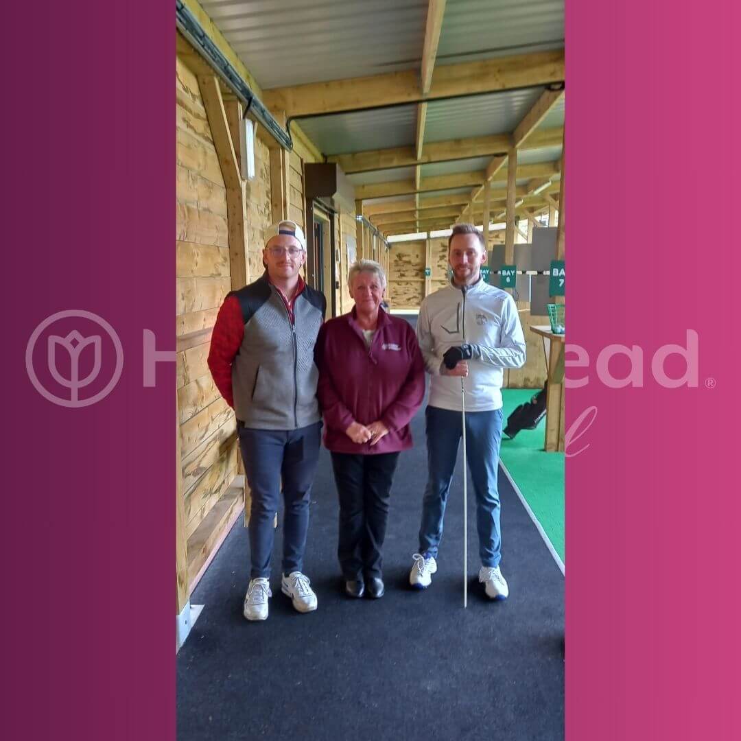 Three people stand side by side in a wooden structure at a golf range, smiling and holding golf clubs. - Home Instead