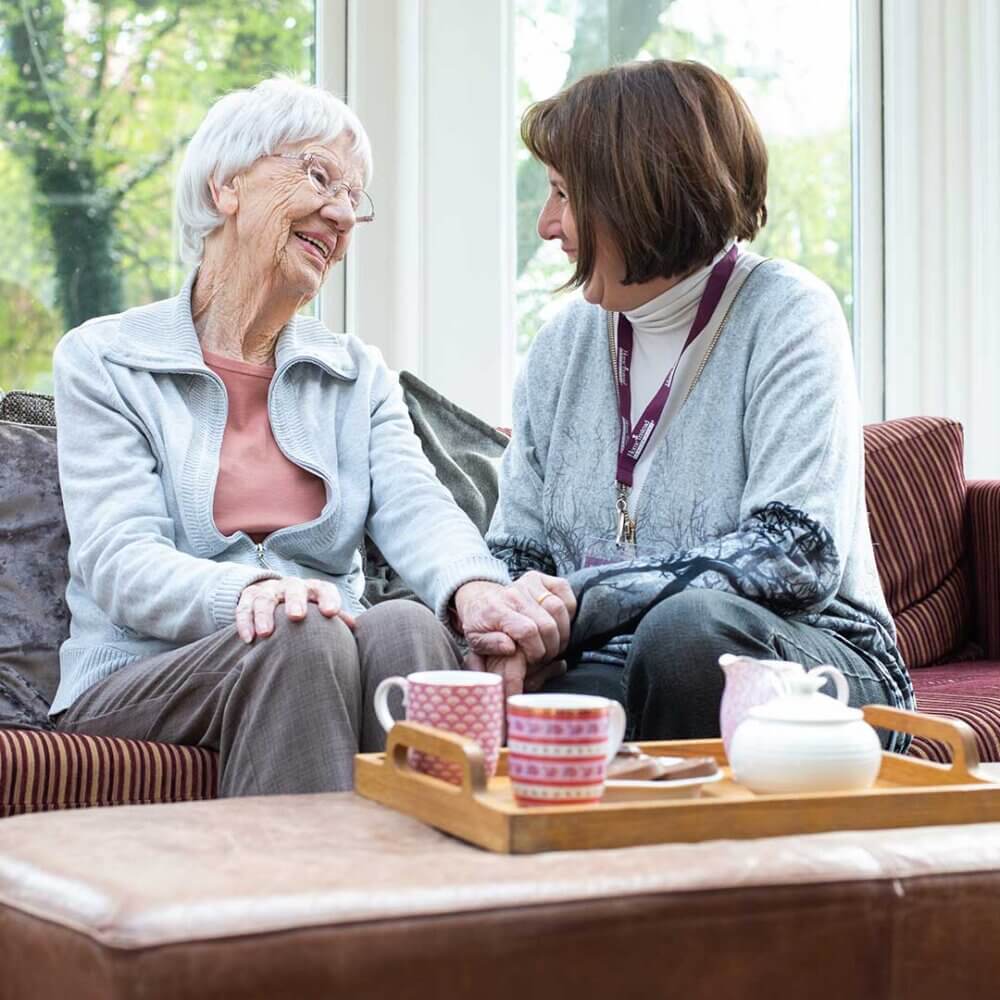 Two women sit on a couch, smiling and holding hands, with tea cups and a teapot on a tray in front of them. - Home Instead