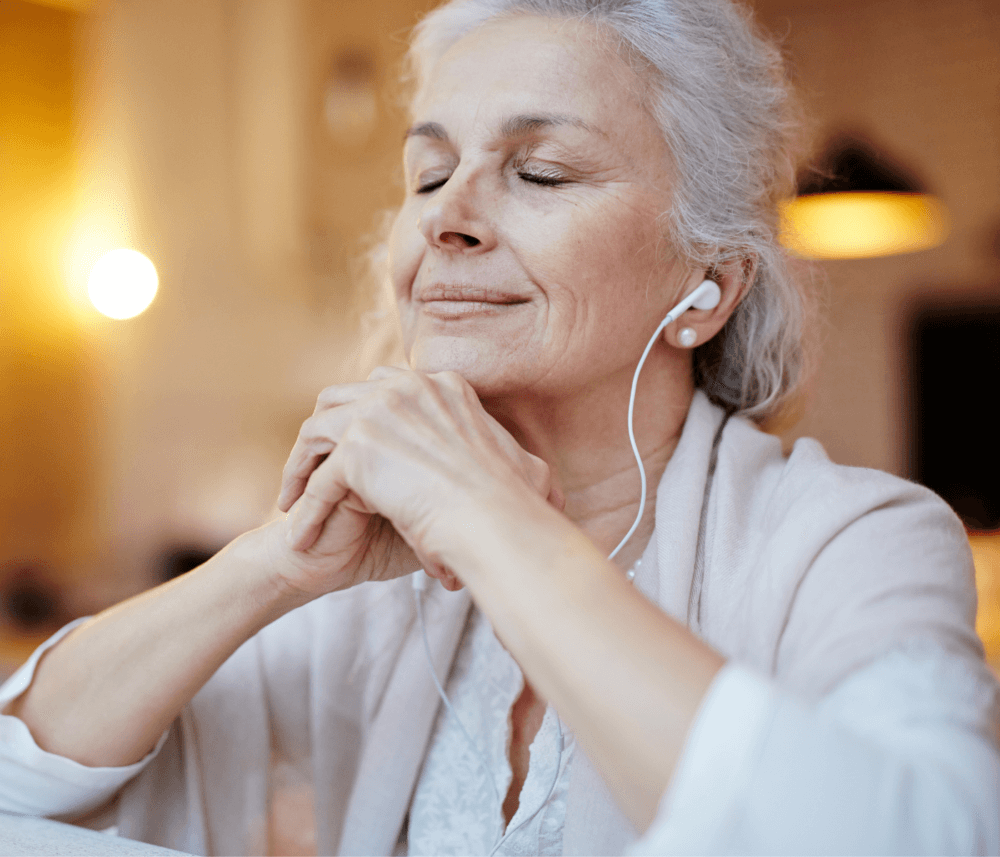 Elderly woman with closed eyes, listening to music through earphones, hands clasped, and smiling peacefully. - Home Instead