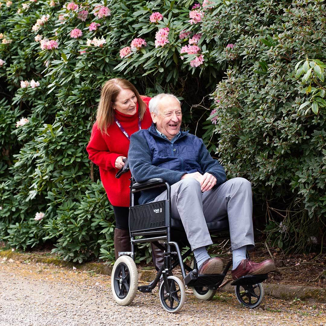 A woman in a red coat pushes an older man in a wheelchair along a path beside blooming plants. Both are smiling. - Home Instead