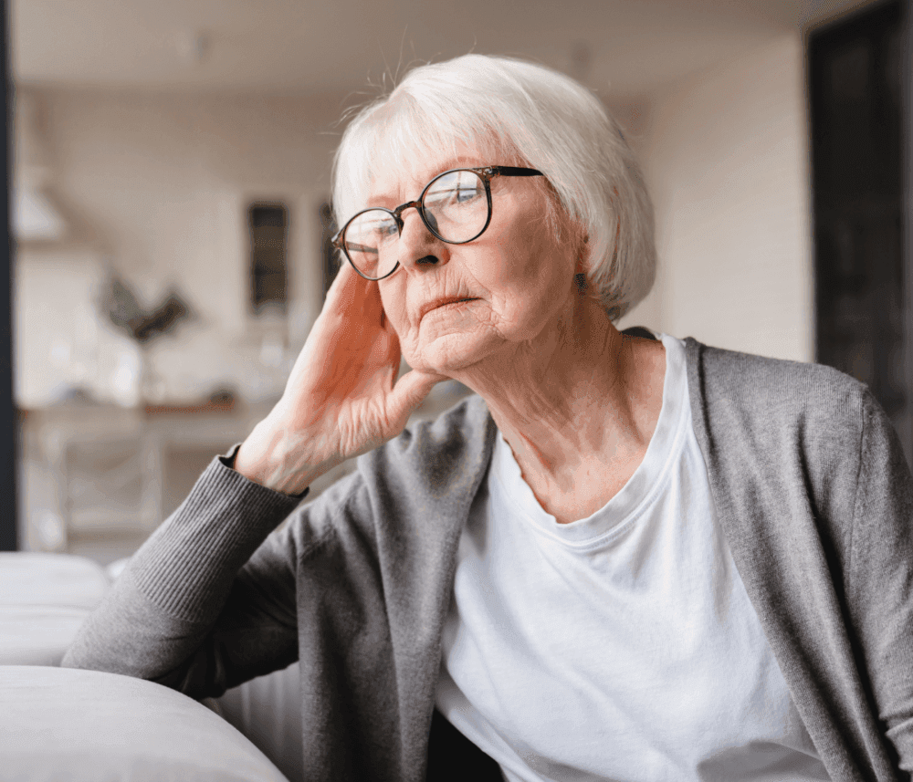 Elderly woman with short white hair and glasses, wearing a grey cardigan, sitting and gazing thoughtfully. - Home Instead