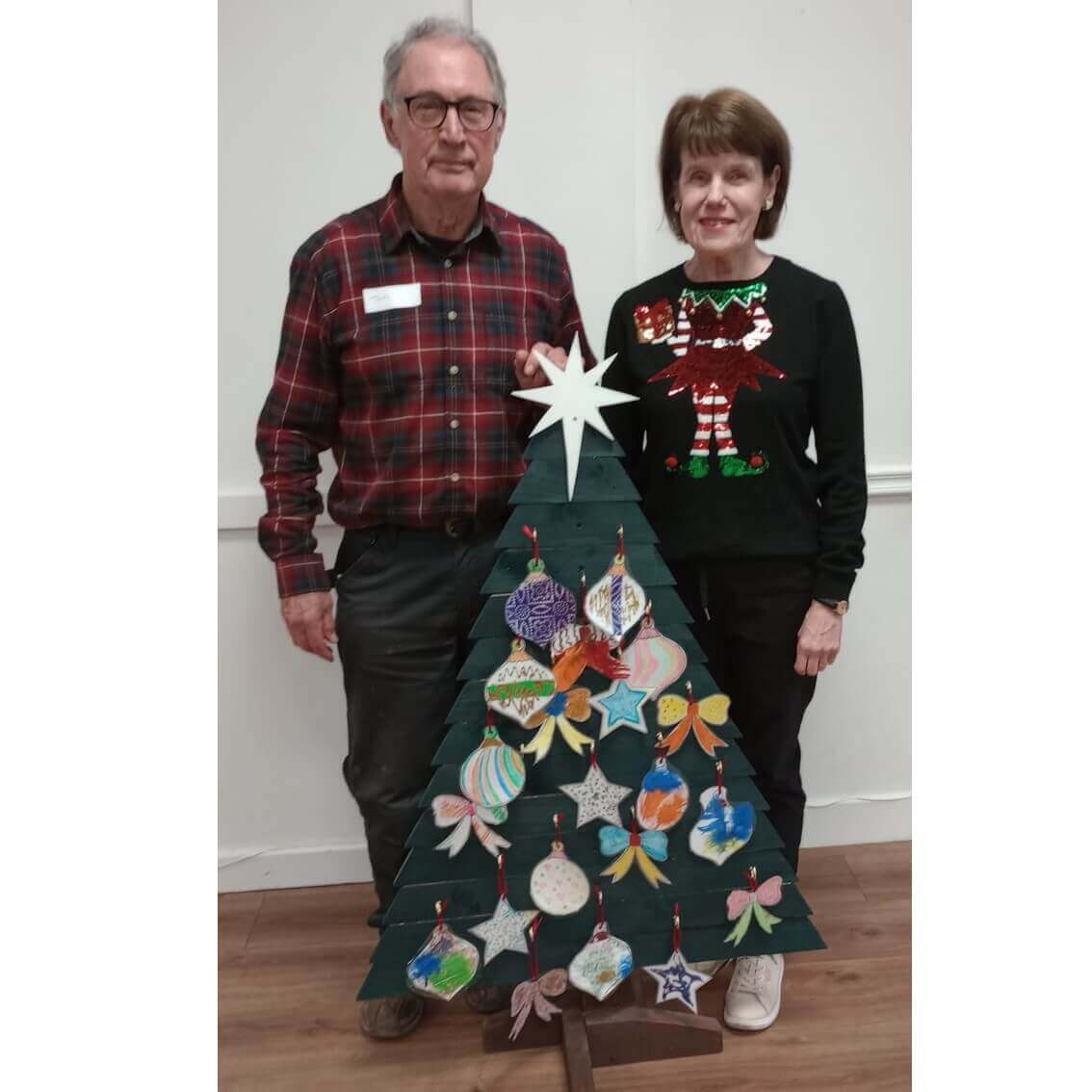 Two people standing beside a wooden Christmas tree adorned with colorful ornaments in a festive setting. - Home Instead