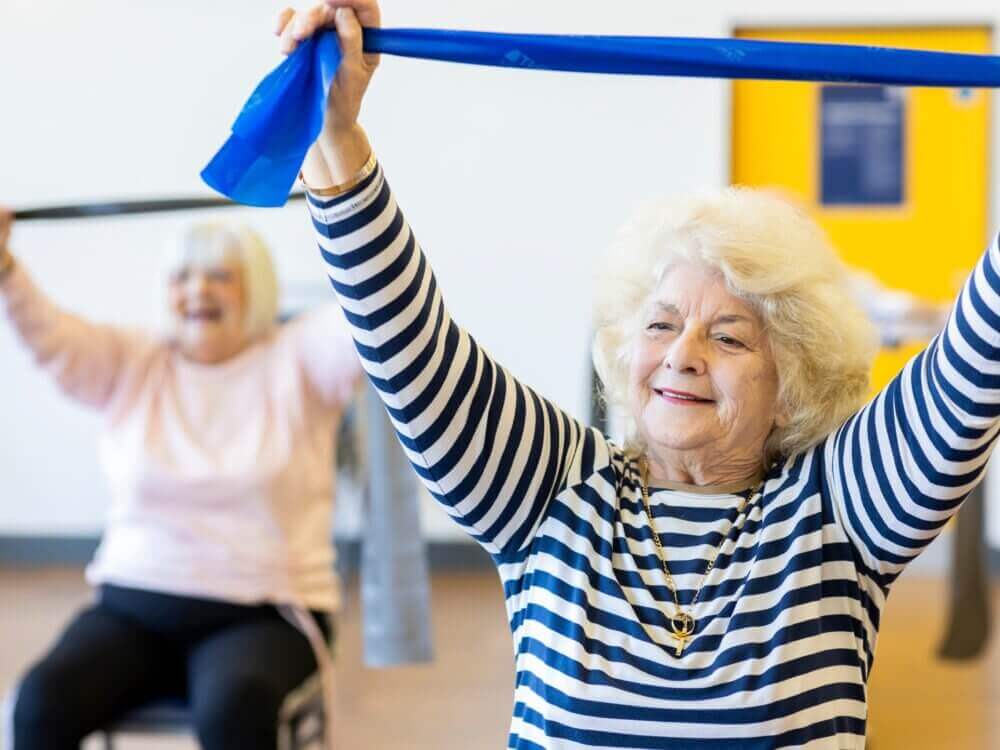 Two elderly women smiling as they stretch exercise bands during a fitness class in a bright room. - Home Instead