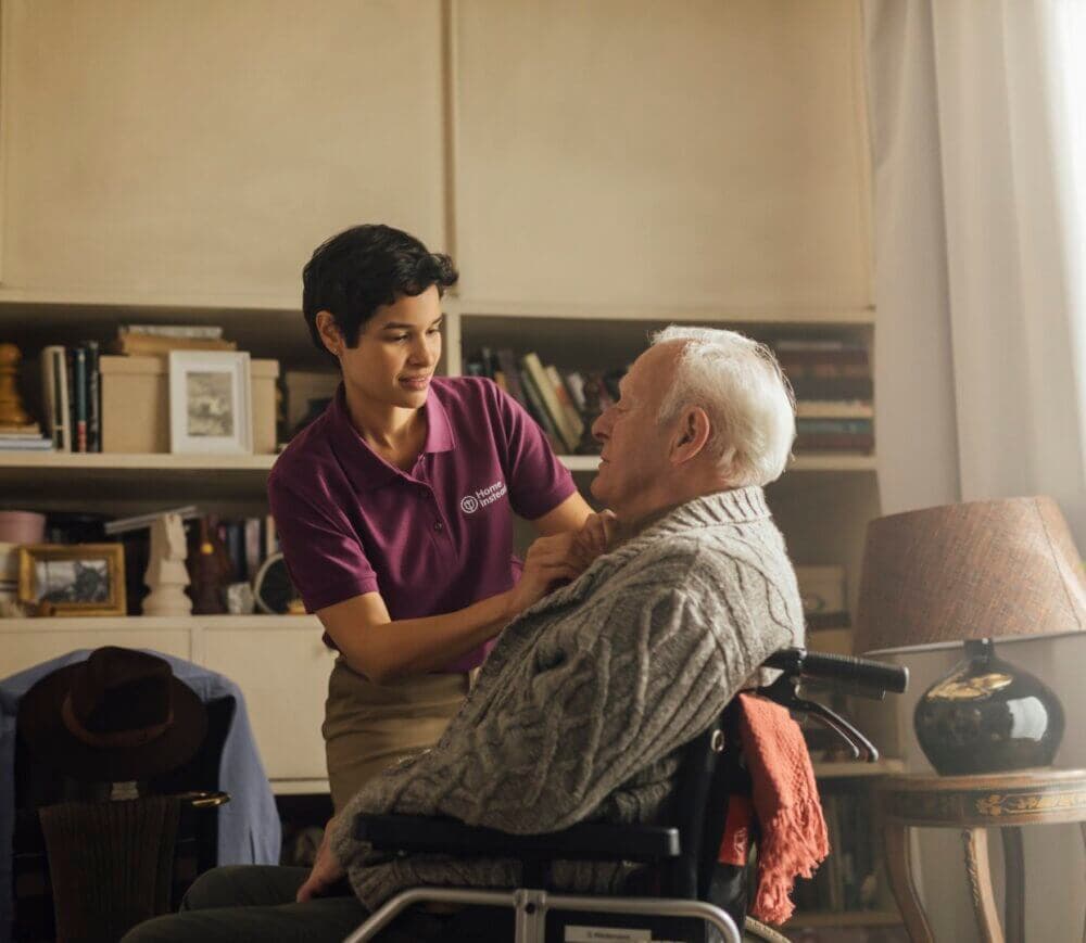 A caregiver in a purple shirt helps an elderly man in a wheelchair button his sweater in a cozy, book-filled room. - Home Instead