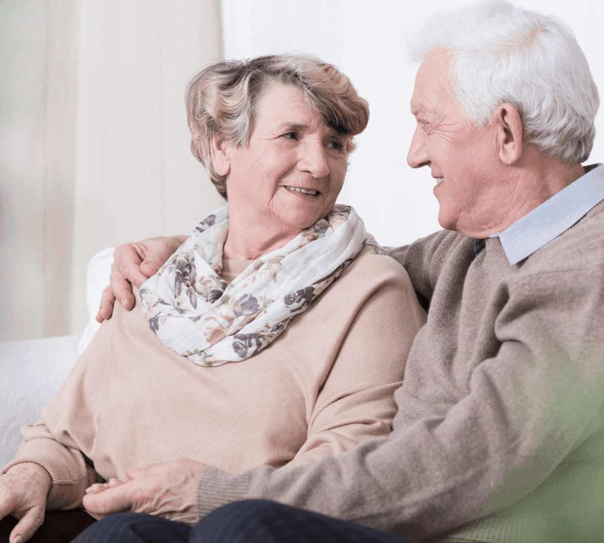 Elderly couple sitting close on a couch, smiling and looking at each other lovingly. - Home Instead