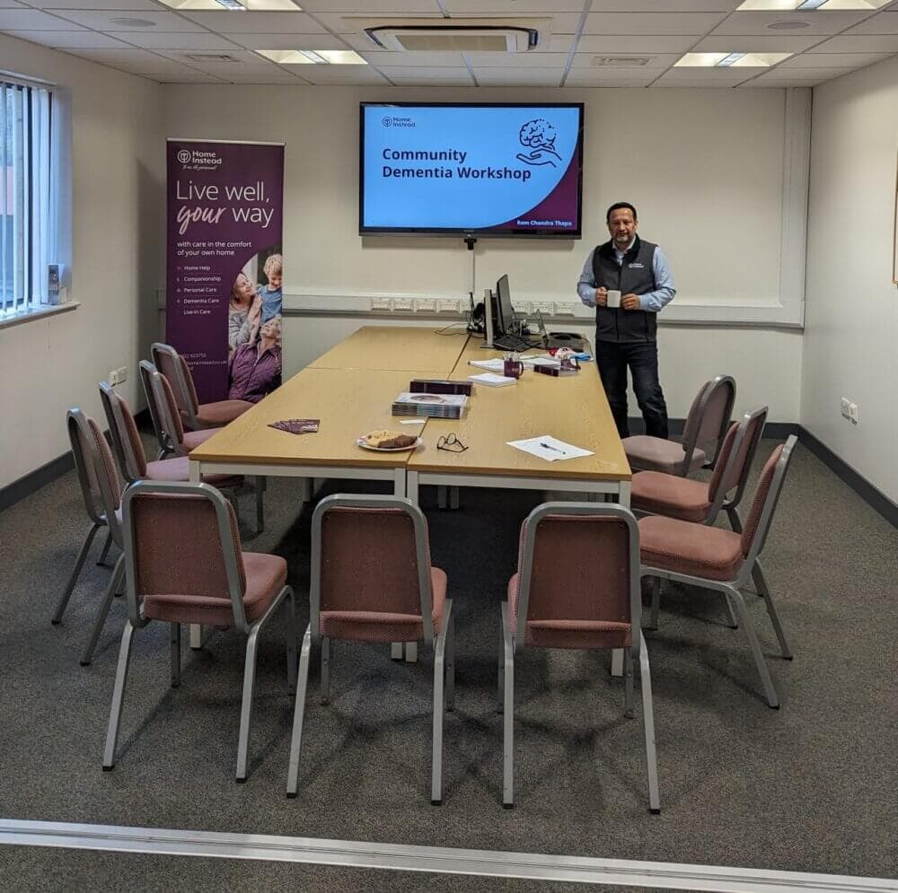A man stands beside a table in a meeting room set up for a Community Dementia Workshop. A screen displays the workshop title. - Home Instead