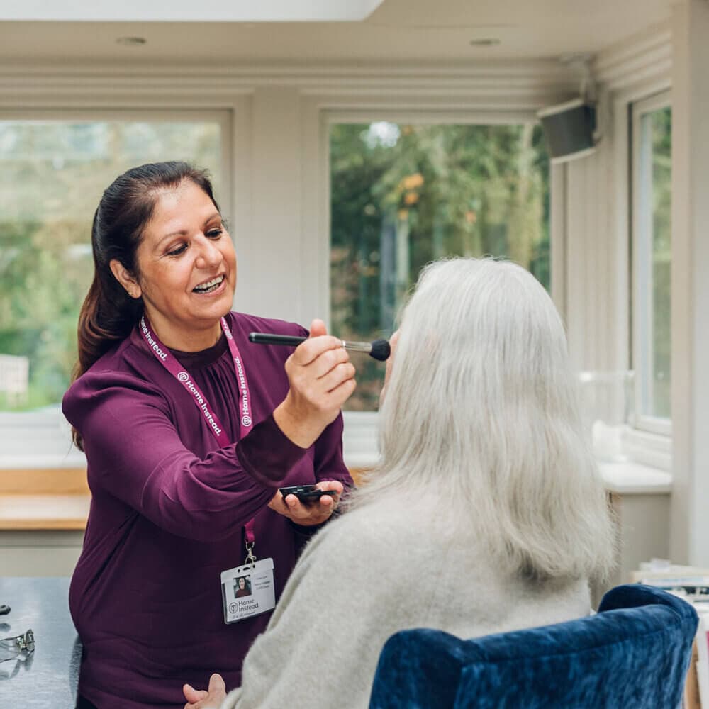 A caregiver smiling while applying makeup to a seated elderly woman in a bright, sunlit room. - Home Instead