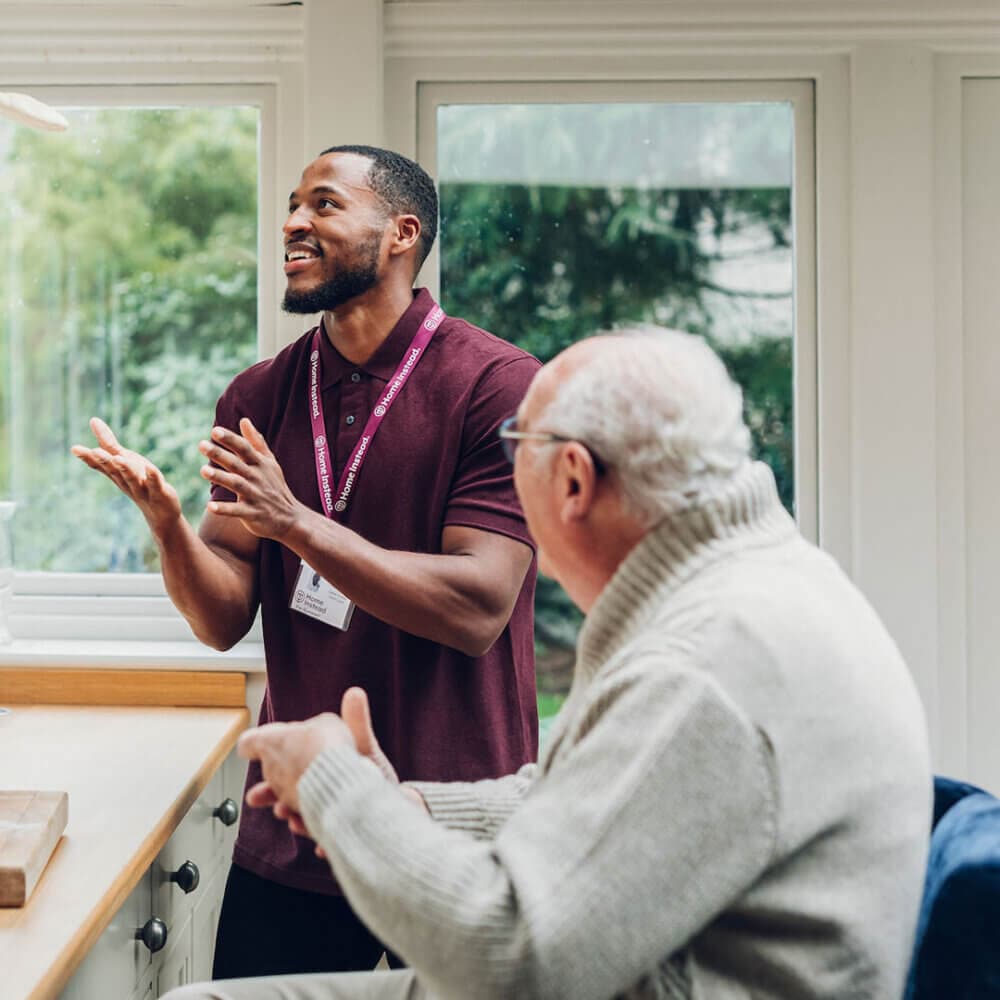 A caregiver with a nametag talks and gestures to an elderly man, seated in a bright room with large windows. - Home Instead