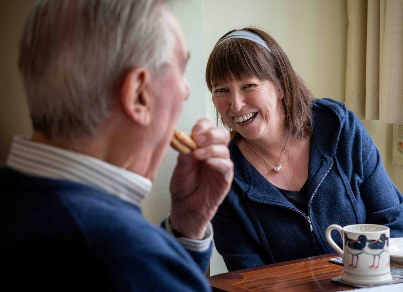 A woman and an elderly man share a laugh while sitting at a table with mugs and plates. - Home Instead