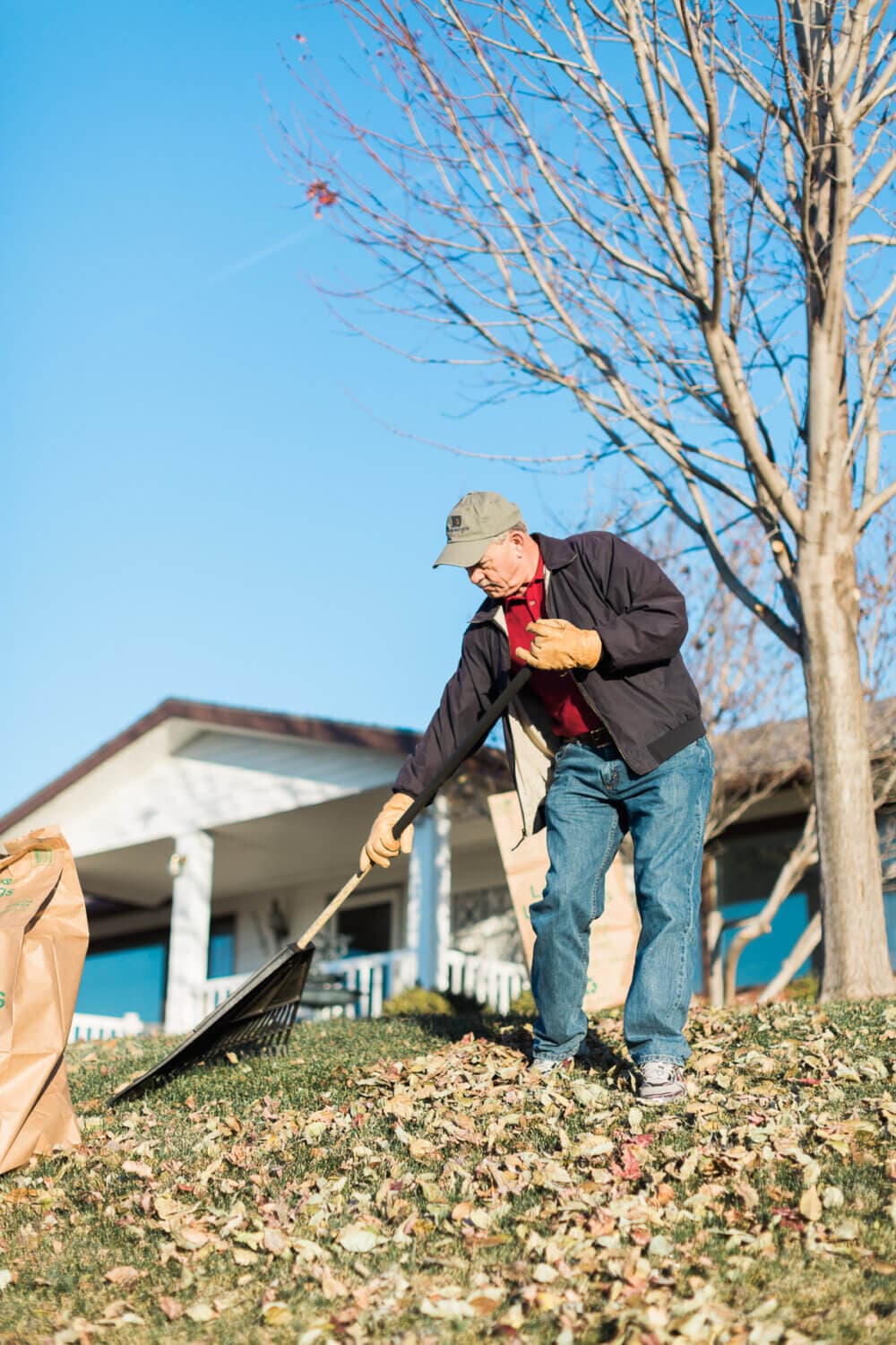Elderly man raking fallen leaves on a sunny day, with a paper yard waste bag nearby. House and tree in the background. - Home Instead