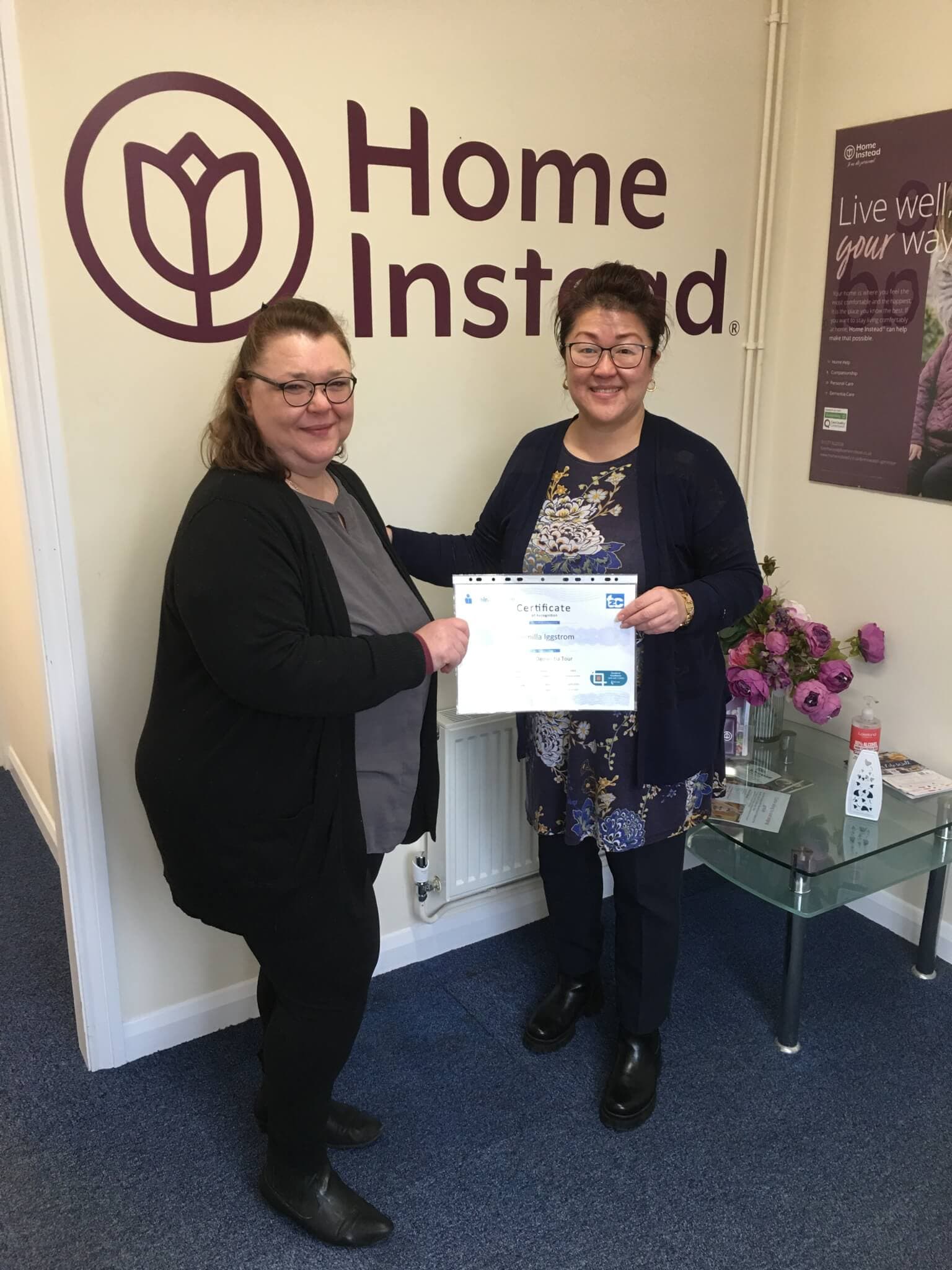 Two women, one holding a certificate, standing in front of a wall sign that reads "Home Instead" with a flower logo. - Home Instead