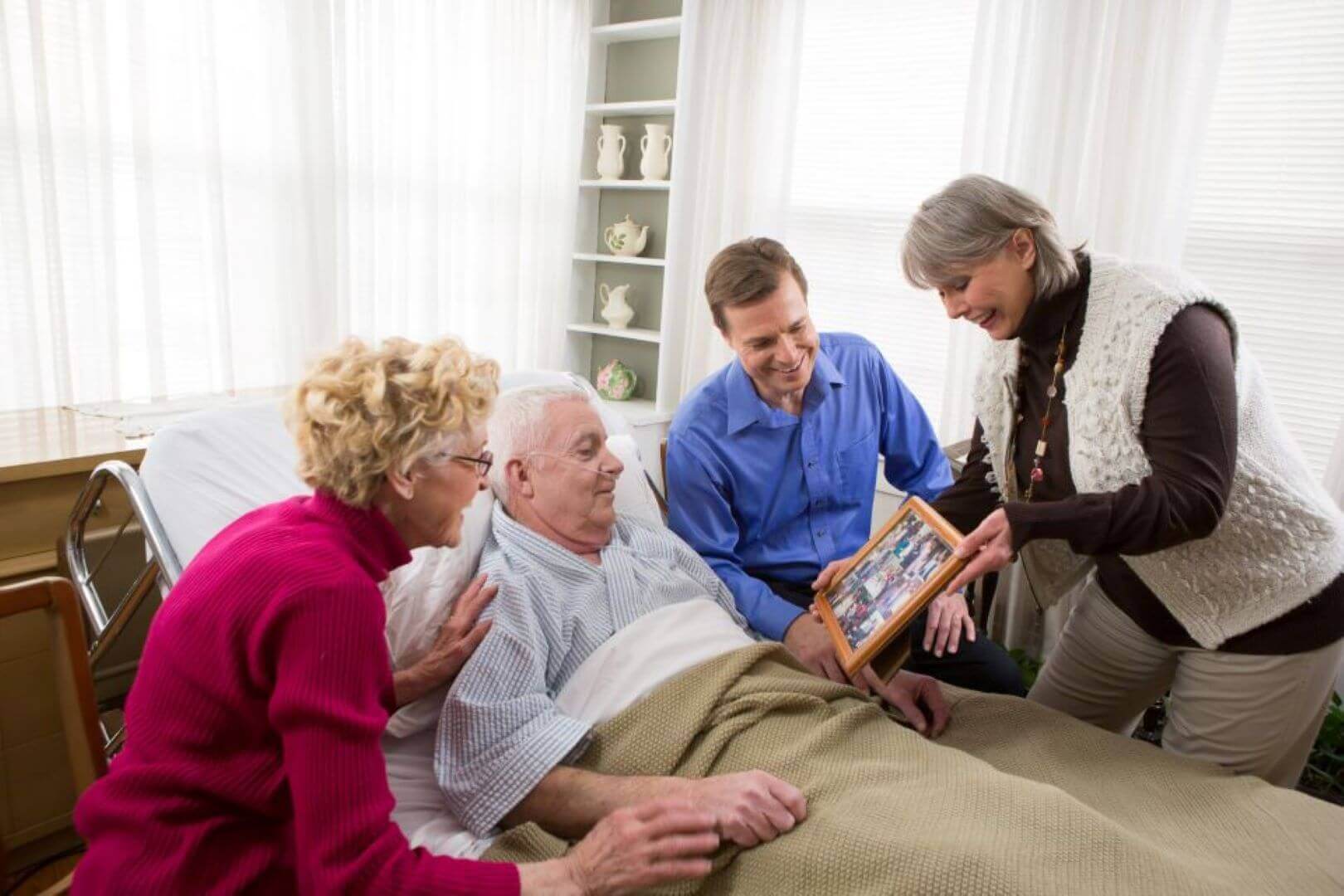 Elderly man in bed, surrounded by two women and a man, looking at a photo frame in a bright room. - Home Instead