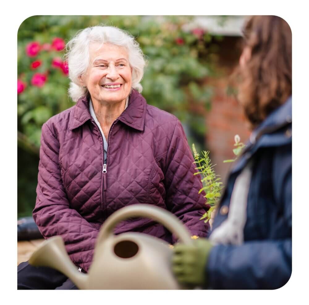 An elderly woman smiles while sitting in a garden, engaging in conversation with another person holding a watering can. - Home Instead