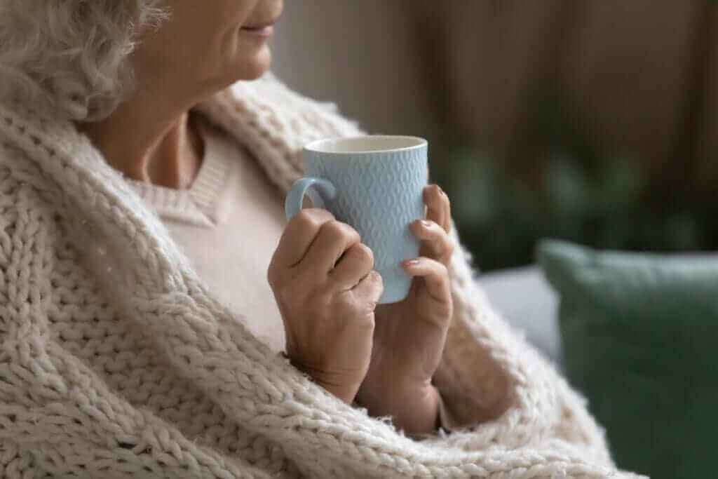 Elderly person wrapped in a blanket holding a blue mug and looking into the distance. - Home Instead