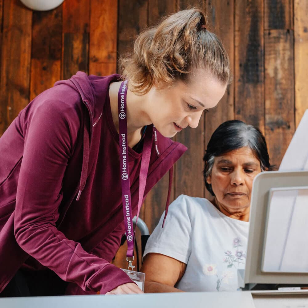 A woman in a maroon jacket helps an older woman with a computer against a wooden wall background. - Home Instead