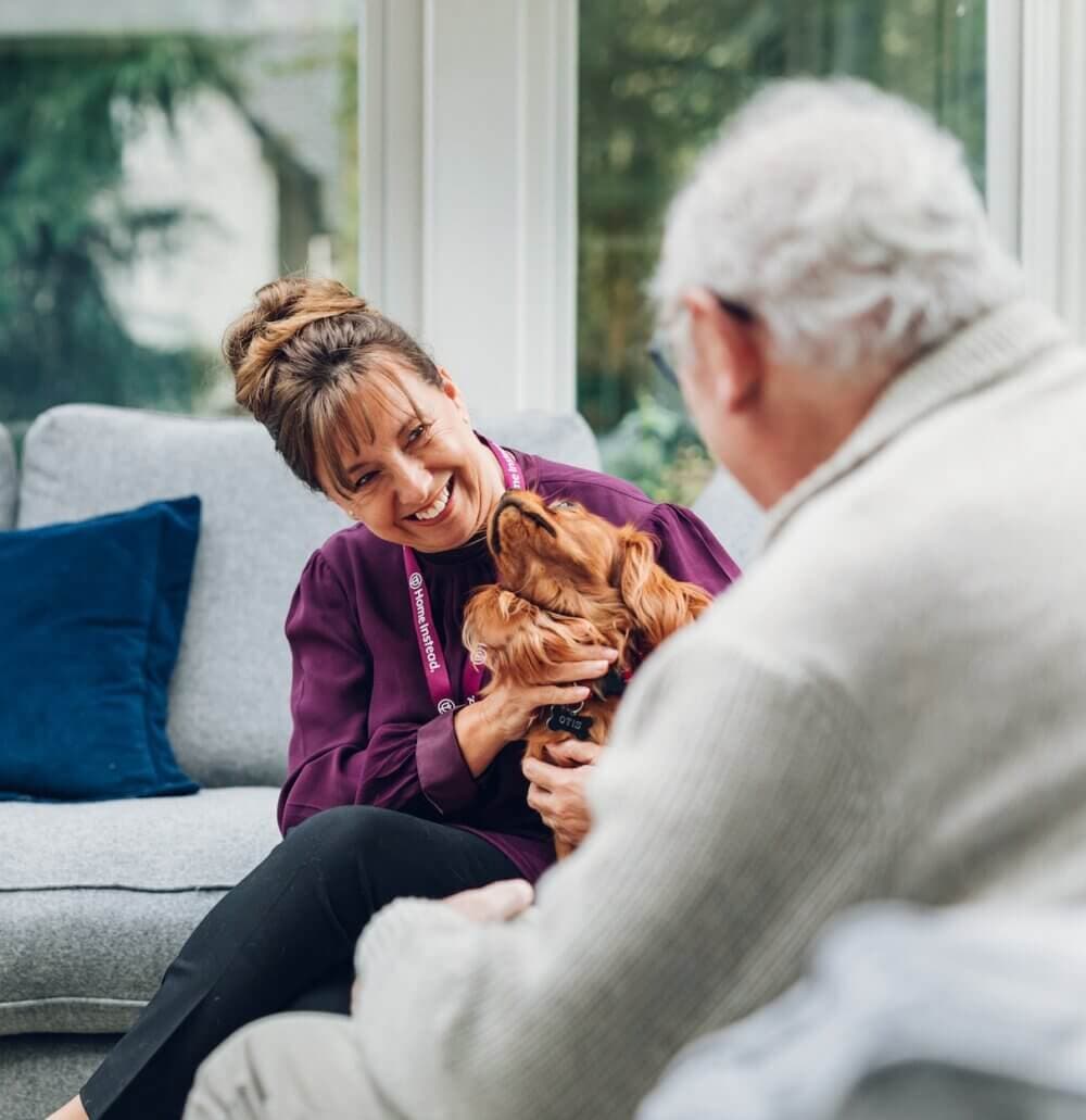 A caregiver and an elderly man smile at each other while the caregiver holds a dog, sitting on a couch indoors. - Home Instead
