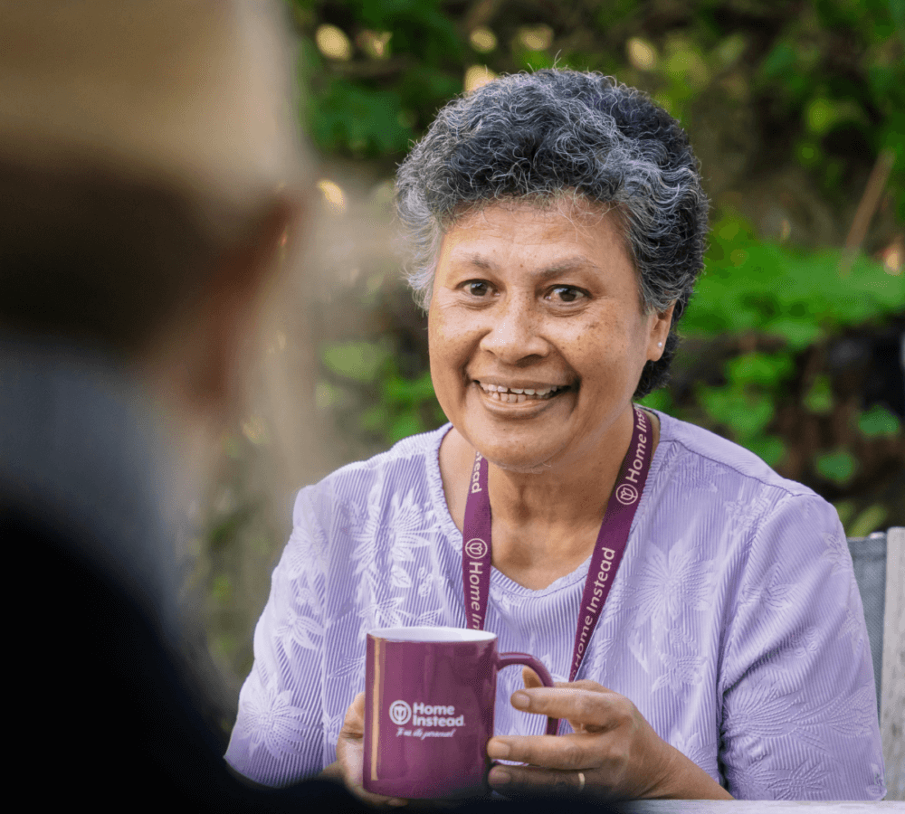 Person with gray hair smiles while holding a Home Instead mug, wearing a purple top and lanyard. Blurred background. - Home Instead