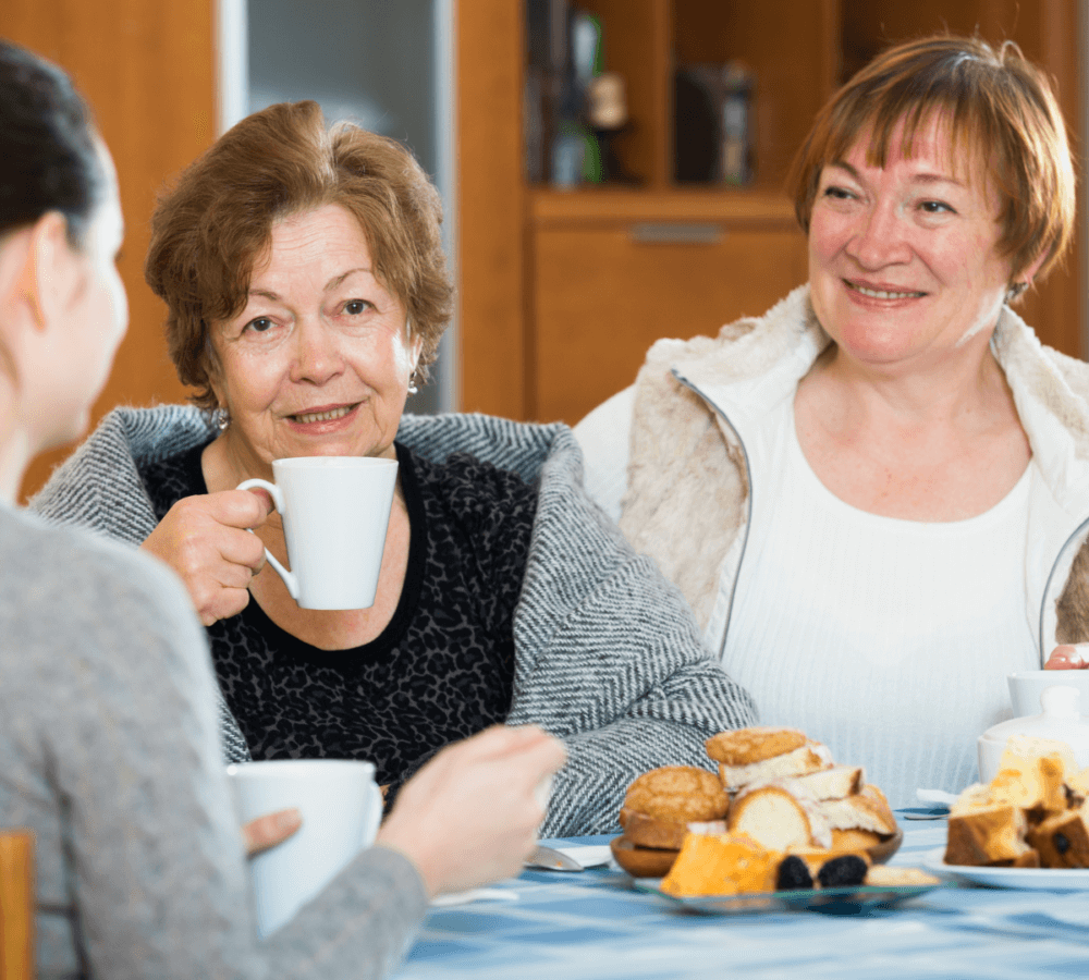 Three women sitting at a table, enjoying hot drinks and pastries, and smiling while engaging in conversation. - Home Instead