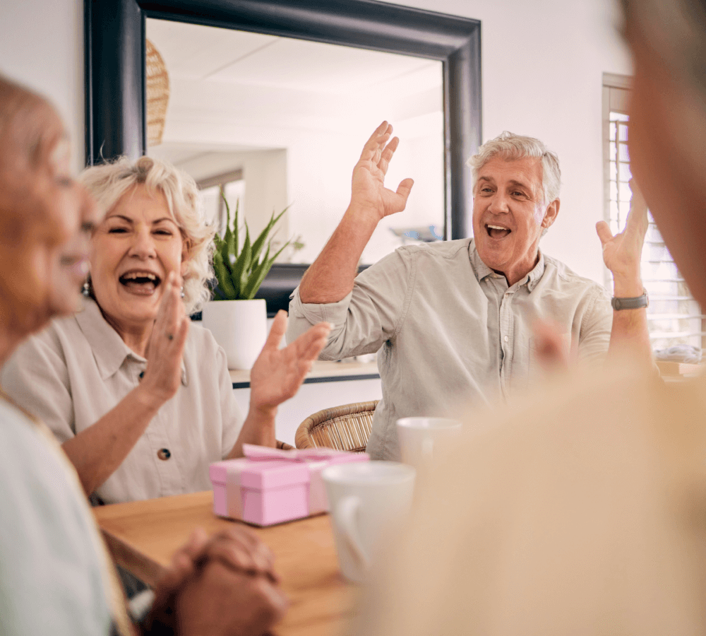 Two older adults are clapping and smiling, seated at a table with pink gift boxes and teacups, reflecting joy. - Home Instead