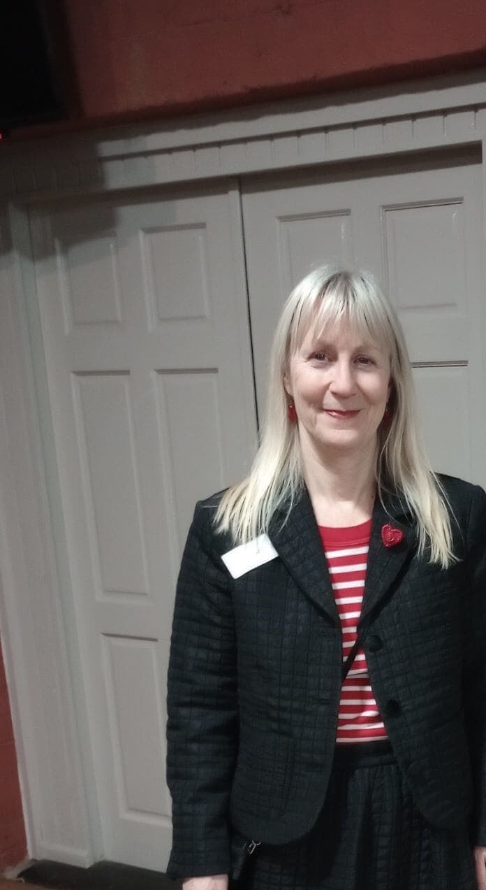 Woman with long blonde hair in a black jacket and red striped shirt stands in front of double doors. - Home Instead