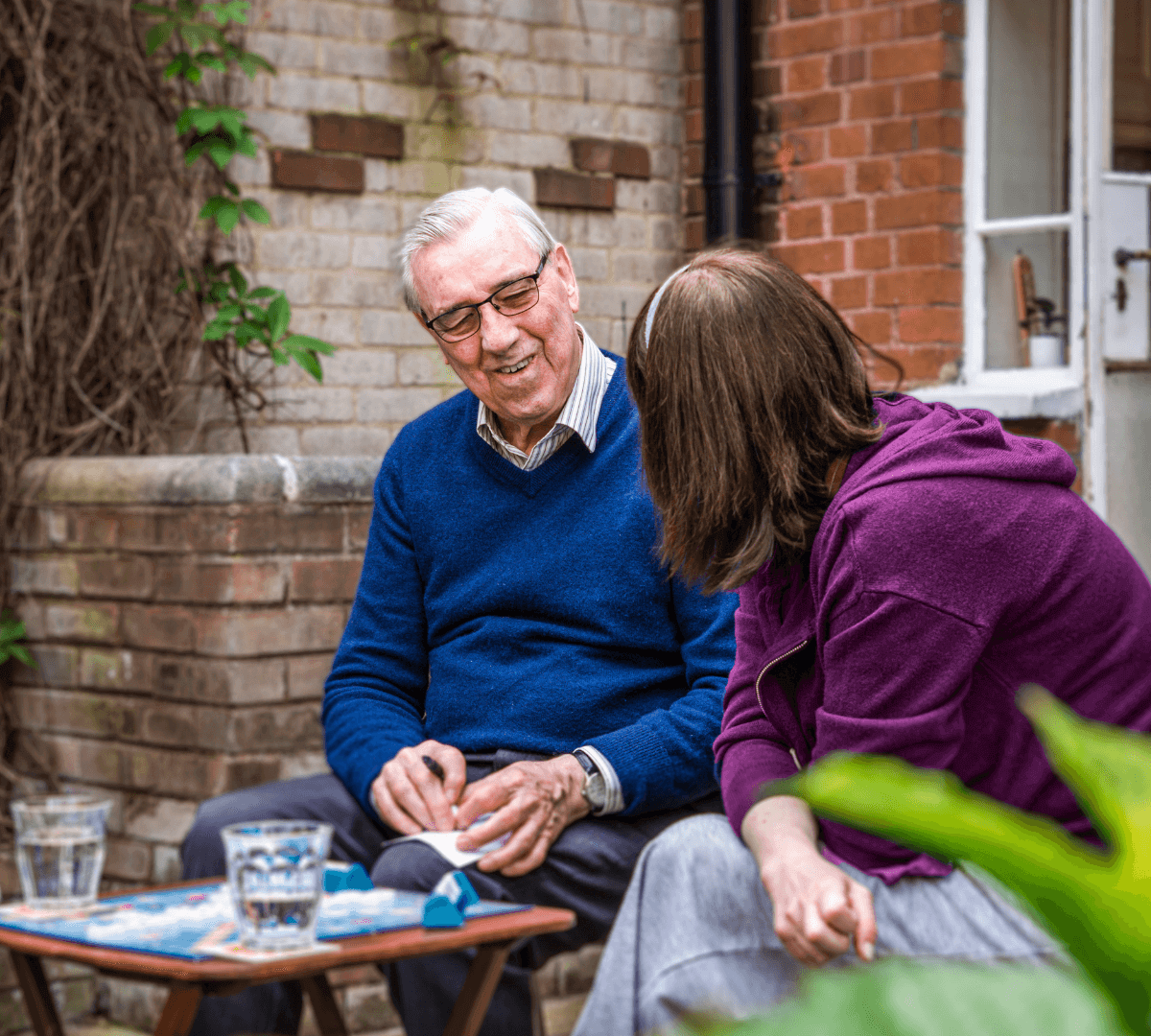 An elderly man and a woman sit outdoors on chairs, engaged in conversation, with drinks on the table in front of them. - Home Instead Southampton