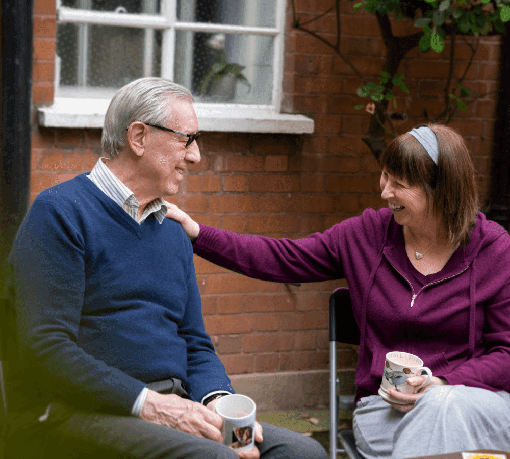An elderly man and a woman smiling and holding mugs while sitting outside a house with a brick wall and a window. - Home Instead Southampton