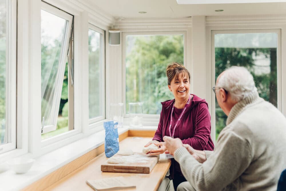 A woman and an elderly man smile and chat while baking together in a bright kitchen with large windows. - Home Instead