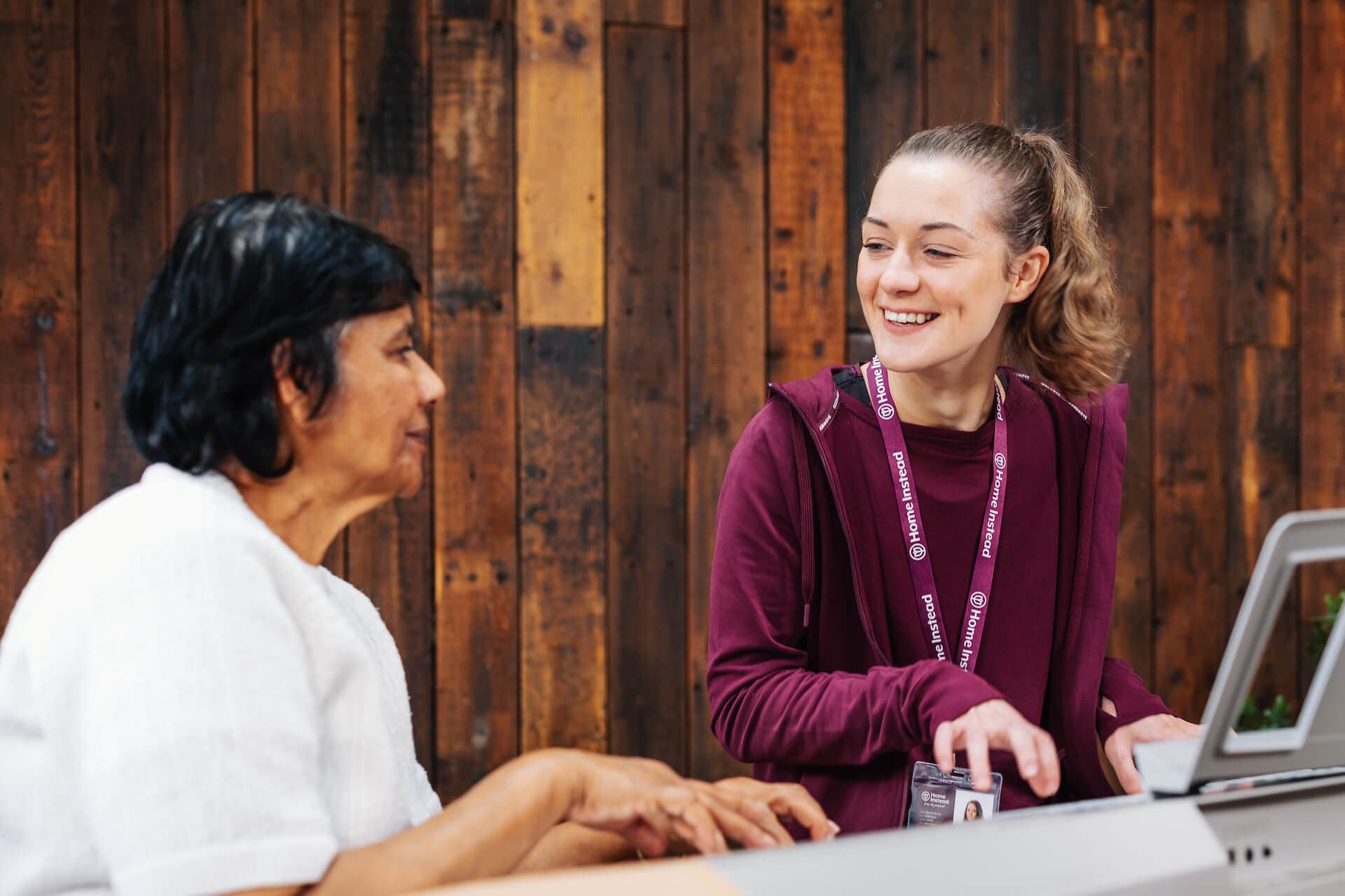A smiling young woman helps an older woman using a tablet, set against a wooden panel background. - Home Instead